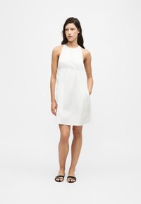 Femme debout portant une robe blanche sans manches au genou avec des poches et des sandales slides noires sur un fond blanc uni.
