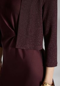 Vestido burdeos con un diseño anudado en la cintura, complementado por una chaqueta corta, texturizada y brillante de mangas tres cuartos.