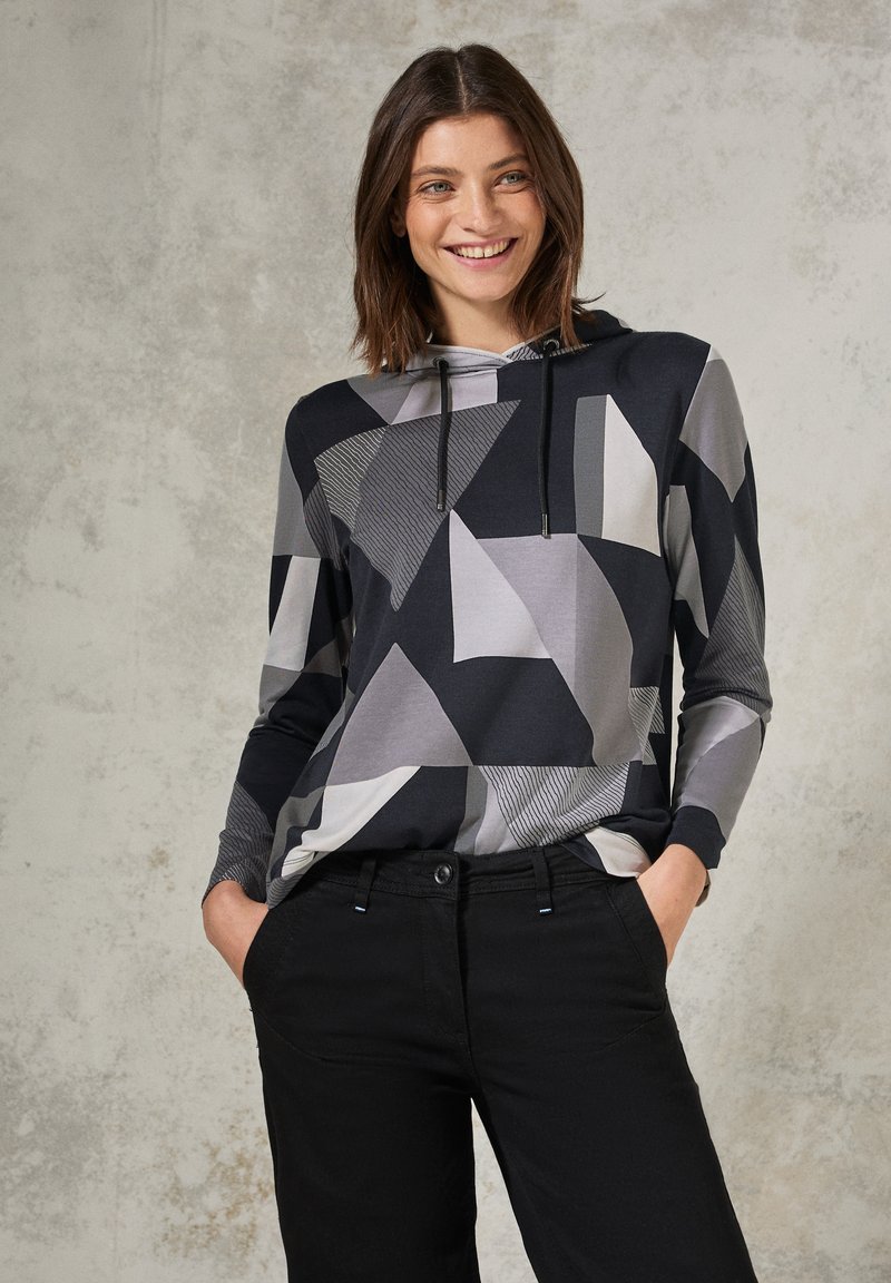 Zwart en grijze hoodie met geometrische patronen en een koord. Gecombineerd met zwarte broek, weergegeven tegen een gestructureerde achtergrond.