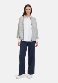 Vrouw staat in een witte blouse, grijze gebreide cape met zakken, marineblauwe wijde broek en witte plateau sneakers.