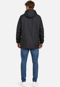 Homme debout de dos, portant une veste noire à capuche, un jean bleu et des baskets blanches sur un fond clair uni.