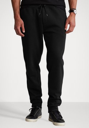 Mand iført sorte joggingbukser med snøre i taljen, sorte sneakers og et armbåndsur, stående mod en ensfarvet baggrund.