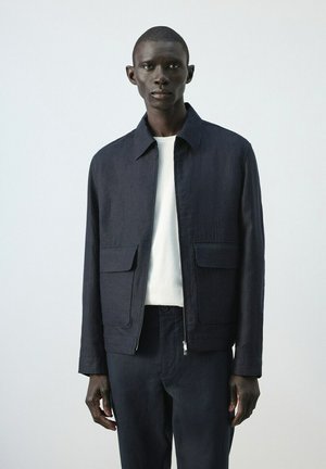 Mężczyzna w ciemnogranatowej kurtce na zamek z klapkowymi kieszeniami, noszący białą koszulę i dopasowane ciemnogranatowe spodnie na jednolitym jasnym tle.