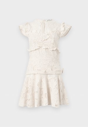 Γυναίκα που φοράει φόρεμα από κρεμ δαντέλα με βολάν στις άκρες, καφέ σανδάλια και στρογγυλά μαύρα γυαλιά ηλίου, στέκεται σε απλό φόντο.