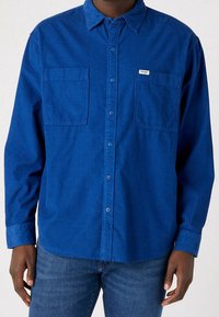 Blå skjorta med knappar gjord av ett texturerat tyg med två bröstfickor, en spetskrage och knäppning vid ärmsluten. Har en liten logotyp.