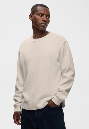 Homme portant une chemise beige clair à manches longues avec texture gaufrée, une main dans la poche d'un pantalon foncé, regardant vers sa gauche.