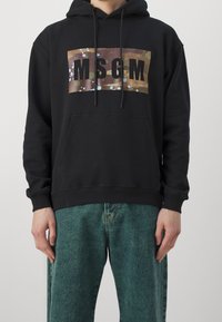 Czarna bluza z kapturem z naszywką z logo w kamuflażowym wzorze z napisem „MSGM” wytłuszczonymi czarnymi literami. Miękka bawełniana tkanina, kieszeń kangurka i ściągacze przy rękawach.
