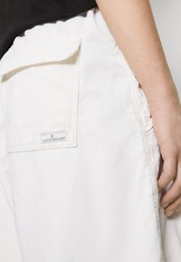 Vita kordflöjtsbyxor har en sidoficka och elastisk midja. De har en texturerad yta med vertikal ribbning och en synlig etikett.