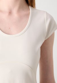 Lichtbeige, getailleerde top met een ronde hals en korte mouwen; gemaakt van glad, rekbaar stof met naaddetails aan de voorkant.