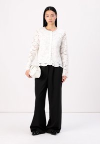 Top lungo a maniche in pizzo bianco con chiusura anteriore a bottoni, abbinato a pantaloni neri larghi e una piccola borsa bianca. Scarponcini neri senza lacci con borchie.