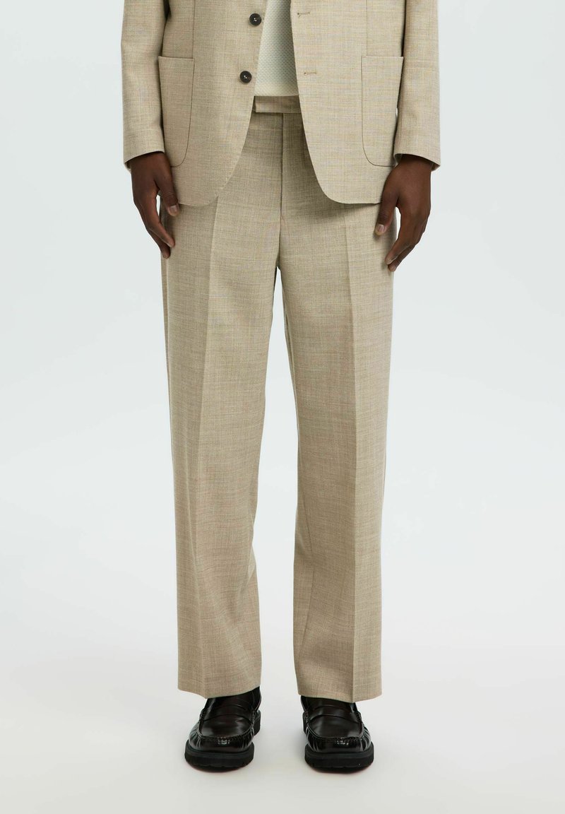 Homme portant un pantalon et une veste de costume beige avec des chaussures en cuir noires, debout devant un fond clair uni.