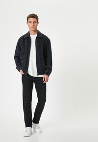 Chaqueta de denim negra con puños elásticos y dos bolsillos frontales, usada sobre una camisa blanca. Pantalones negros ajustados y zapatillas blancas completan el atuendo.