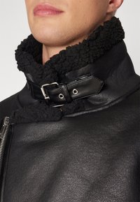 Sort læderjakke med en tekstureret finish og en sort, blød shearling-krave, der lukkes med en metalspænde og stropdetalje.