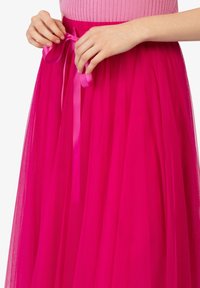 Ruce zavazuj�ící jasně růžovou saténovou stuhu na živě růžové tylové sukni, kterou nosí s růžovým žebrovaným topem na bílém pozadí.