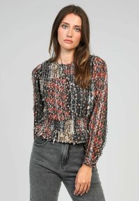 Femme aux longs cheveux bruns portant un haut fleuri multicolore texturé et un jean gris taille haute, se tenant contre un fond clair.