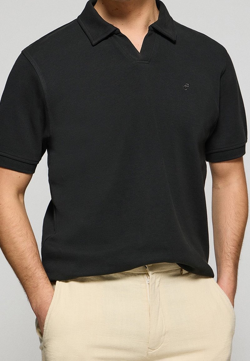 Homme portant un polo noir uni à manches courtes rentré dans un pantalon beige, les mains dans les poches, sur un fond uni.