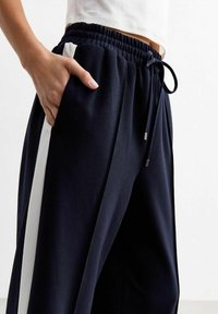 Marineblauwe broek van een gestructureerde stof, elastische tailleband, trekkoord en een witte zijstreep. Zichtbare zijzak aan de linkerkant.