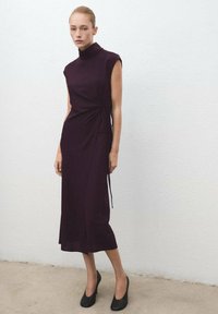 Mujer de pie contra una pared blanca, vestida con un vestido midi sin mangas de color púrpura oscuro con detalle de lazo lateral y tacones altos negros.