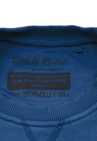 Blå bomullssweatshirt med ribbad halsringning, med svarta tryckta etiketter som anger märke, material och skötselråd.