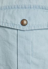 Ljust blå denimtyg med detalj av en framficka, utrustad med en rund metallknapp och synlig söm.