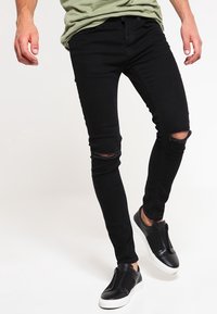 Jean skinny en denim noir avec des déchirures au genou, associés à des chaussures noires à enfiler. Le tissu a une texture lisse et une coupe ajustée.