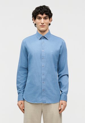 Jeune homme aux cheveux bouclés et foncés portant une chemise en denim bleu clair boutonnée et un pantalon beige, debout devant un fond clair uni.