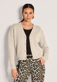 Frau in beiger, geknöpfter Strickjacke über schwarzem Oberteil mit goldenen Creolen und Leopardenmuster-Hose, posiert vor hellem Hintergrund.