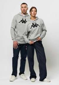 Grijze hoodies met een groot zwart logo op de borst, gecombineerd met donkerblauwe jeans en witte sneakers. Ontspannen pasvorm en zachte textuur.