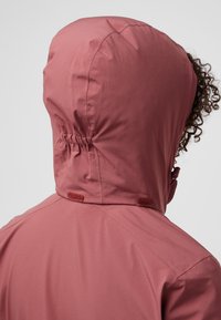 Veste imperméable rose clair avec une capuche ajustée, des fronces élastiques et des coutures plates. Elle présente des fermetures Velcro au niveau du cou et une texture lisse.