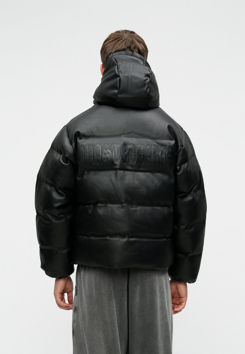 Črna puhovka iz sijočega materiala, z visokim ovratnikom in embosiranim detajlom na hrbtni strani. Kvadratna oblika in prešita tekstura.