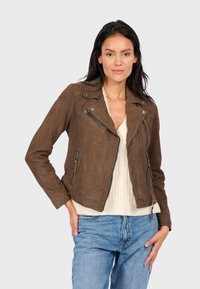 Hnědá semišová motocyklová bunda se stříbrnými zipy, přiléhavý střih a šikmé kapsy. Nositá přes světle krémový top a modré džíny.