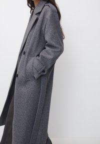 Abrigo de lana gris con una silueta larga y recta, grandes botones, bolsillos de parche y cuello con muesca. Textura suave y diseño minimalista.