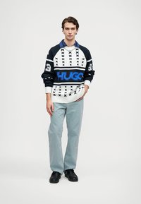 Pulóver fekete, fehér és kék színekben; geometrikus minta; nagy "HUGO" logó; világoskék texturált nadrággal és fekete cipővel párosítva.