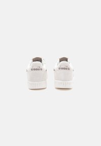 White suede sneakers with a textured finish, featuring a light brown accent on the heel and logo detail in metallic gray.