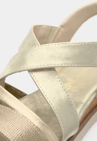 Sandale à brides croisées en beige métallique avec une semelle intérieure texturée et un talon en bois. Comprend des brides synthétiques lisses et des détails de couture contrastants.