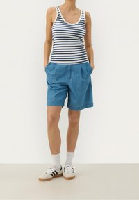 Shorts de mezclilla azul con pliegues y bolsillos laterales, combinados con una camiseta sin mangas a rayas en azul marino y blanco. Usados con calcetines blancos y zapatillas deportivas.