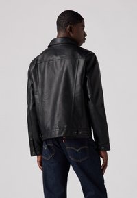 Veste en cuir noir avec un col classique, couture visible et fermeture à boutons-pression, assortie à un jean en denim foncé avec des accents jaunes.