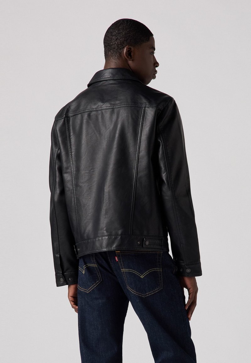 Veste en cuir noir avec un col classique, couture visible et fermeture à boutons-pression, assortie à un jean en denim foncé avec des accents jaunes.