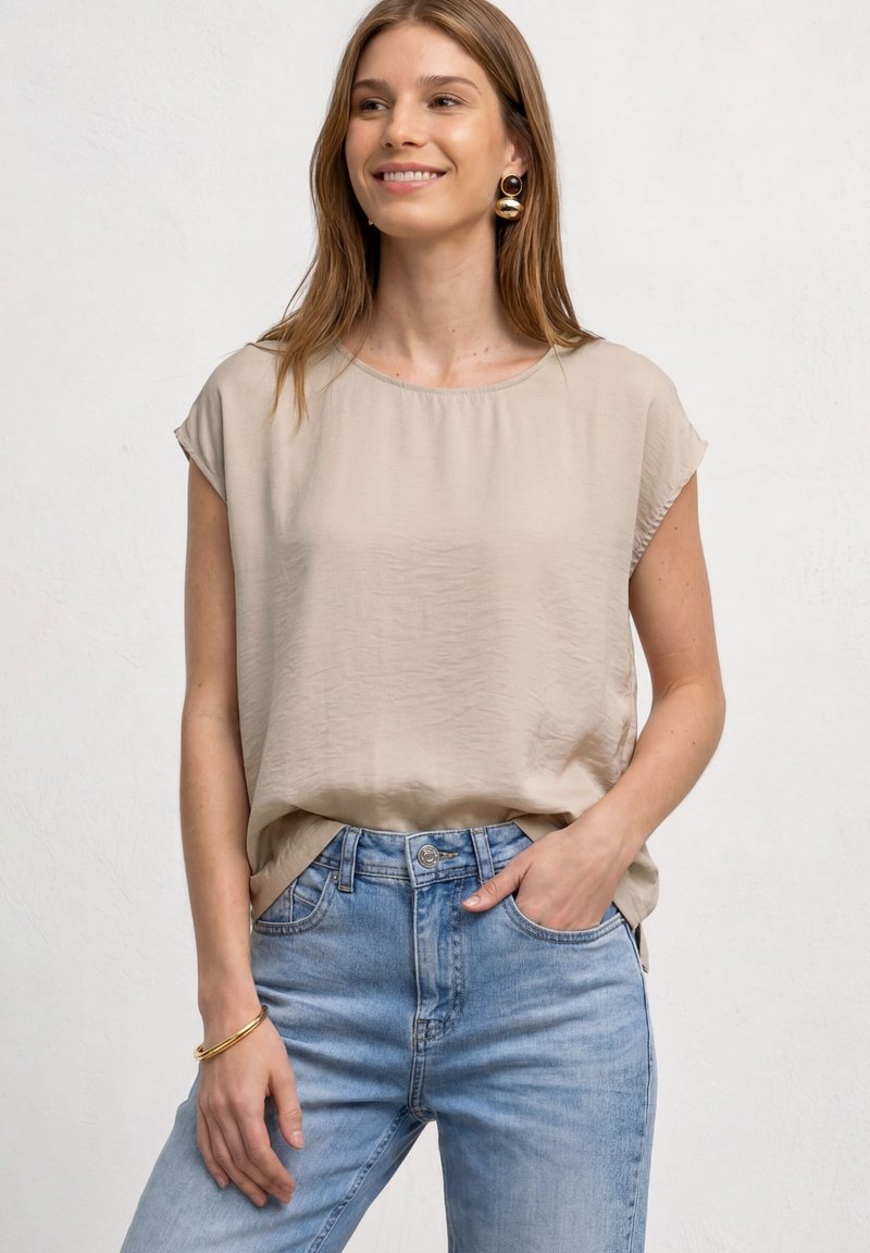 Femme souriante portant un chemisier beige à manches courtes, un jean bleu clair, de grandes boucles d'oreilles rondes en or et un bracelet en or, une main dans la poche.