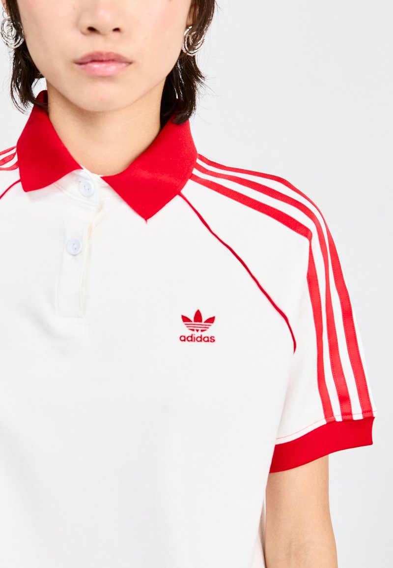Biała koszulka polo z czerwonym kołnierzykiem i czerwonymi paskami na rękawach. Posiada małe czerwone logo Adidas na piersi. Materiał wydaje się być fakturowany.