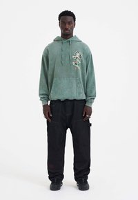 Mintgroene hoodie met een voorzak, geborduurde rozendetails. Gecombineerd met losse zwarte cargo broeken met zijzakken en subtiele branding.