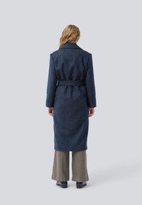 Mujer con largo cabello rubio que lleva un abrigo de lana azul oscuro con cinturón, pantalones de pierna ancha beige y zapatos negros, de pie sobre un fondo gris claro.