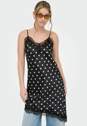 Kvinde iført sort satin natkjole med hvide polkaprikker og blonder, kombineret med lyseblå jeans og solbriller med ravfarvet glas.