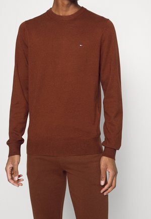 Homme portant un pull ajusté à manches longues couleur rouille et un pantalon assorti, debout devant un fond clair uni.