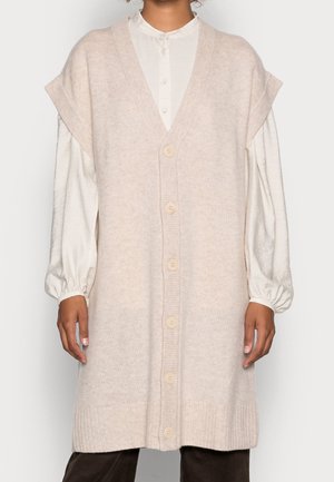 Personne portant un long cardigan sans manches en maille beige avec des boutons, par-dessus un chemisier blanc à manches longues et un pantalon foncé.