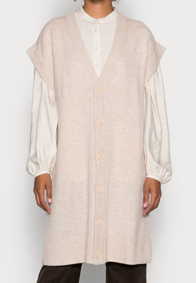 Personne portant un long cardigan sans manches en maille beige avec des boutons, par-dessus un chemisier blanc à manches longues et un pantalon foncé.