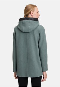 Grøn tekstureret frakke med en aftagelig sort hætte, der har en lige pasform og mellemlang længde, båret over sorte bukser.