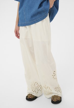 Persona con pantalones sueltos blancos bordados, combinados con sandalias color canela y una blusa de mezclilla azul, de pie frente a un fondo blanco liso.