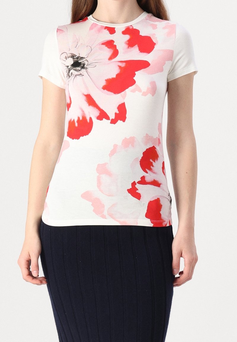 T-shirt blanc à manches courtes avec un imprimé floral rouge et rose. Tissu doux, encolure ronde, coupe ajustée. Associé à une jupe côtelée foncée.