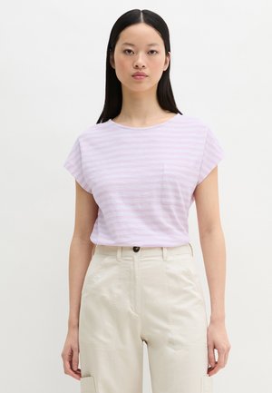 Femme aux longs cheveux noirs portant un t-shirt rayé violet clair avec une poche poitrine et un pantalon beige taille haute sur un fond uni.